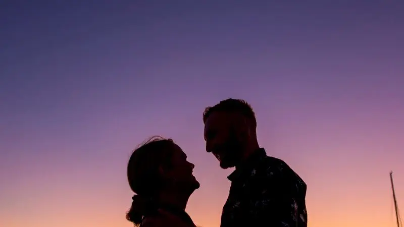 Silhouetted couple embraces on a sunset jetty after a Sundowner Sunset Cruise, with glowing sailing boats and scenic hills in the background.