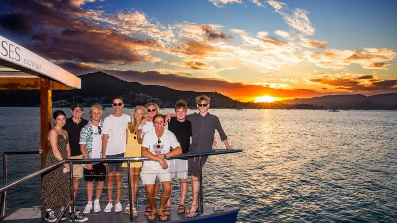 Nine friends enjoy a Sundowner Sunset Cruise, smiling as they pose with scenic mountain views and sparkling water in the background.
