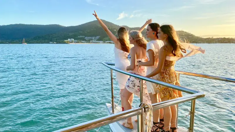 Four women in stylish dresses smile on a sunset cruise boat, enjoying panoramic sea views and scenic mountains in the background.