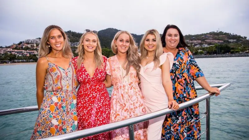 Five women in vibrant dresses enjoy a Sundowner Sunset Cruise on a boat, smiling with scenic water and hills in the glowing background.