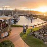 Vibrant sunset over marina with moored sailboats, people walking, and Sundowner Cruise & Dine at Sorrentos Restaurant visible on left.