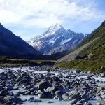 Scenic rocky river winds through lush green valley with snow-capped Mount Cook under clear blue sky, ideal for Queenstown tours.