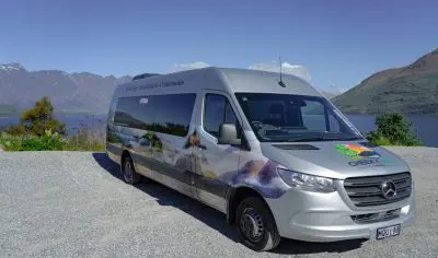 Silver tour van parked beside picturesque lake near Queenstown, snow-capped mountains, clear blue sky—scenic New Zealand travel scene.