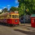 Vintage red tram and classic phone box on a sunlit Christchurch street, framed by leafy trees and iconic historic architecture.