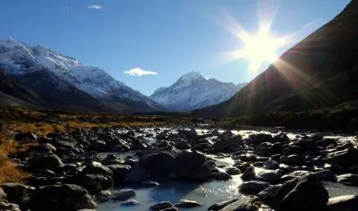 Scenic rocky riverbed with snow-capped peaks near Mount Cook, sunlit under a vibrant blue sky, perfect for nature photography enthusiasts.