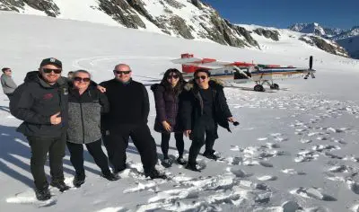 A small tour group poses on snowy terrain, with dramatic mountain peaks and a scenic small aeroplane in the bright sunlight behind them.
