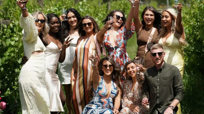 Stylish friends toast with wine glasses at a Yarra Valley winery tour, smiling and enjoying a sunny day in scenic vineyard surroundings.