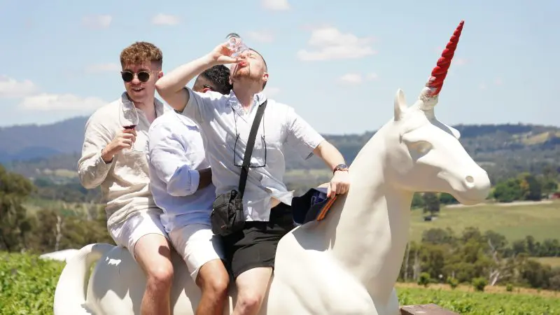 Three men in summer outfits sit atop a white unicorn statue, savouring wine and relaxing on a scenic Yarra Valley winery tour.