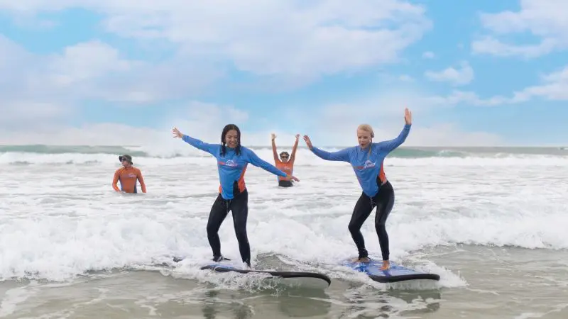 Two women in wetsuits riding small waves during a 2-hour beginner surf lesson for ages 13+, learning surfing techniques on boards.