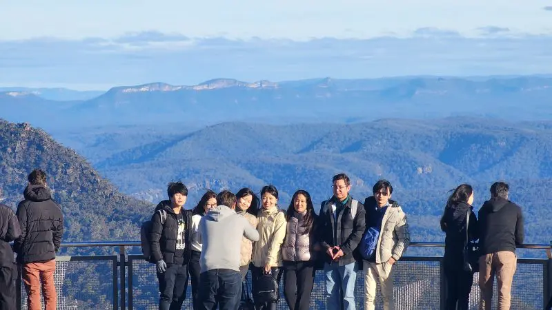 A smiling group enjoys panoramic Blue Mountains views at a scenic lookout on a guided waterfall walk and social lunch tour experience.