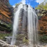 Experience a majestic, slender waterfall tumbling down rocky cliffs on a Blue Mountains Day Tour Waterfall Walk beneath a scenic sky.