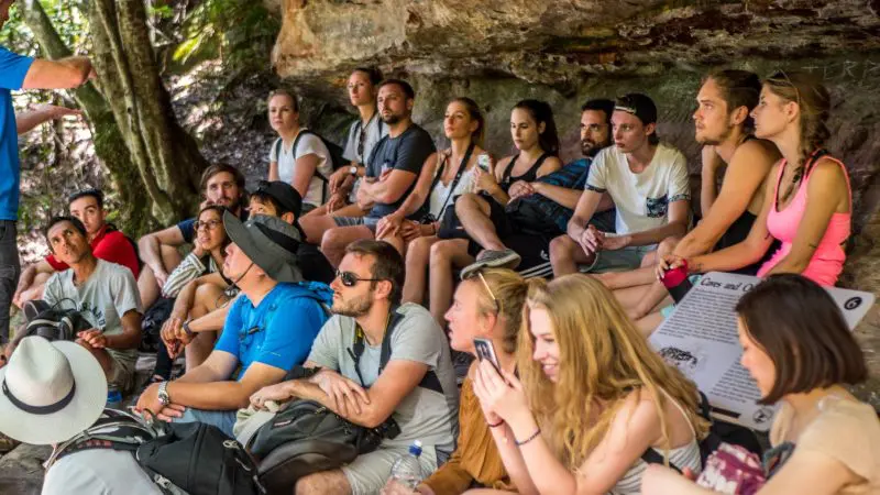 Tourists gather under a scenic Blue Mountains rock ledge on a 1 Day Wildlife Park Trip, engaging attentively with their expert guide.