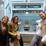 Three women smiling and gesturing to the floor of a spacious, empty hall on a Canberra day trip from Sydney, highlighting local sights.