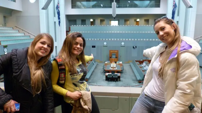 Three women smiling and gesturing to the floor of a spacious, empty hall on a Canberra day trip from Sydney, highlighting local sights.