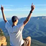 Traveller sits on scenic Blue Mountains cliff edge, arms uplifted, savouring panoramic views during all-inclusive signature day tour.