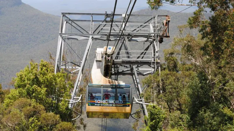 A yellow Scenic World cable car soars above lush forest, showcasing breathtaking Blue Mountains views and Lyrebird Pass scenery.