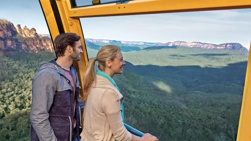 Man and woman admire panoramic mountain and forest views from Lyrebird Pass cable car with expansive windows for optimal sightseeing.