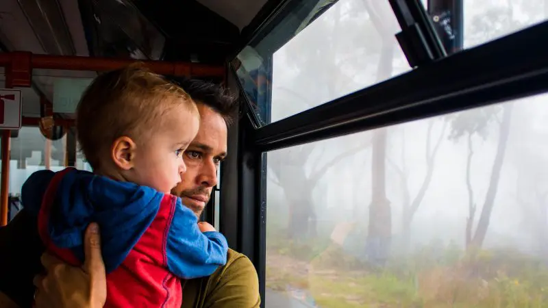 A father cradling his child looks out of a bus window on the Boomerang Pass One Hour Sightseeing Tour, enjoying scenic city views.