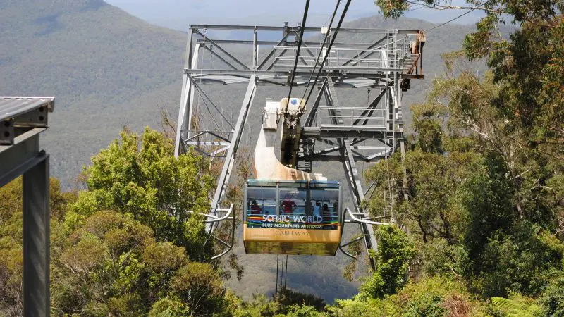 Bright yellow Scenic World cable car soars over lush treetops in Kookaburra Pass with Blue Mountains panorama in the background.