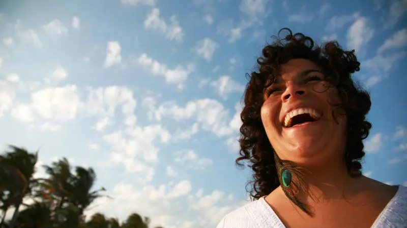 Curly-haired woman laughs joyfully outdoors, enjoying the Hinterland Magic Glow-worm Experience beneath vibrant blue skies.