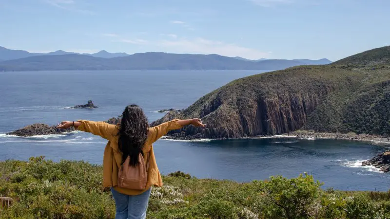 A joyful woman with outstretched arms enjoys stunning ocean cliffs and lush greenery on a Bruny Island Day Tour in Tasmania.