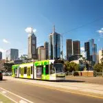 Green and white Melbourne tram travels down a sunlit street with city skyline in the background, highlighting Ultimate Melbourne Adventure.