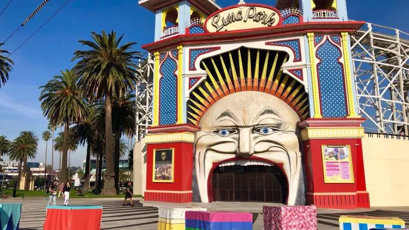 Iconic Luna Park entrance with a giant smiling face, lush palm trees, and vibrant cube seats welcomes visitors to Melbourne’s top attractions.