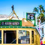 Iconic vintage yellow tram featuring a St Kilda Beach sign, framed by swaying palm trees and clear blue Melbourne skies, epitomising adventure.