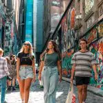 Three fashionable young adults explore a vibrant graffiti alley in Melbourne, wearing trendy summer outfits and sunglasses, soaking up city vibes.