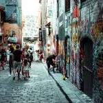 Tourists stroll down a vibrant Melbourne alleyway filled with colourful graffiti, photographing unique street art on their city adventure.