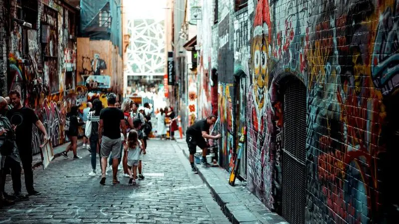Tourists stroll down a vibrant Melbourne alleyway filled with colourful graffiti, photographing unique street art on their city adventure.