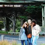 Three smiling women posing at the iconic Great Ocean Road sign with lush trees and markers in the background during a 2-day tour.