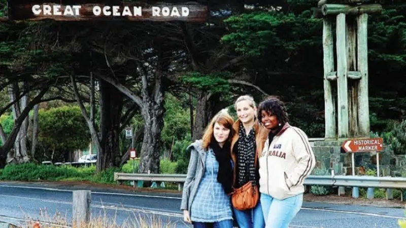 Three smiling women posing at the iconic Great Ocean Road sign with lush trees and markers in the background during a 2-day tour.