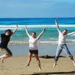 Three people jumping with joy on a sandy beach during the Ultimate 2 Day Great Ocean Road Tour, ocean waves and blue sky in view.