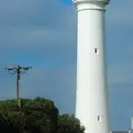 Three tourists stand on a scenic path beside a towering white lighthouse on the Ultimate 2-Day Great Ocean Road Tour in Australia.