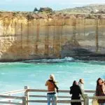 Tourists stand on a scenic wooden lookout platform overlooking dramatic coastal cliffs and crystal-clear turquoise waters on the Great Ocean Road.