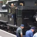 A man waves from a vintage black steam train in the scenic Dandenong Ranges, whilst three uniformed workers sit at the front.
