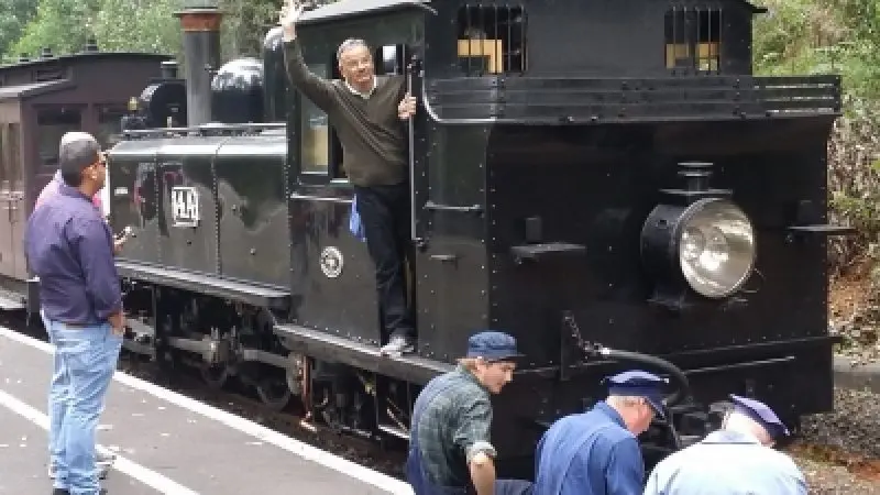 A man waves from a vintage black steam train in the scenic Dandenong Ranges, whilst three uniformed workers sit at the front.