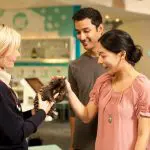 A smiling couple gently touch an echidna held by a woman indoors at Healesville Wildlife Sanctuary, experiencing unique wildlife.
