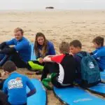 Children and two adults in blue shirts sit on surfboards on a sandy beach, ready for their Learn to Surf Tour surfing lesson.