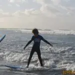Two children at the Learn to Surf Tour: one balances on a surfboard in shallow water, while another stands nearby holding a board.