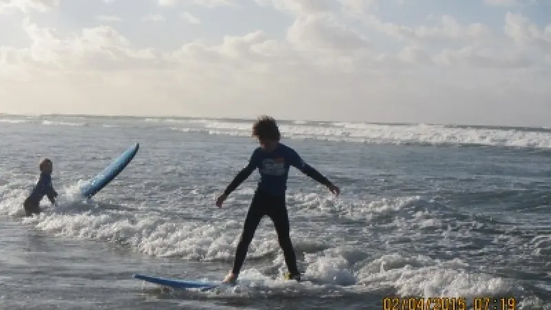 Two children at the Learn to Surf Tour: one balances on a surfboard in shallow water, while another stands nearby holding a board.
