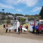 Surfers in wetsuits holding surfboards assemble by the Learn To Surf Tour trailer on a lush, green seaside grassy area.