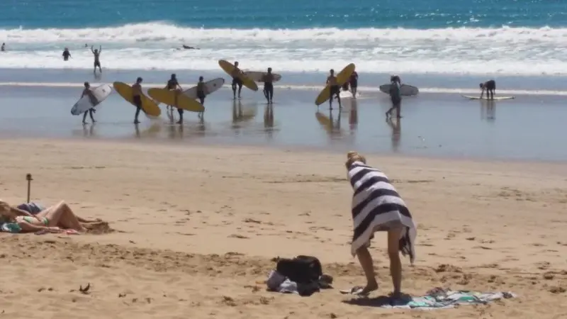 Surfers with boards walk along the ocean shoreline while a Learn to Surf Tour guest, wrapped in a towel, stands on the sandy beach.