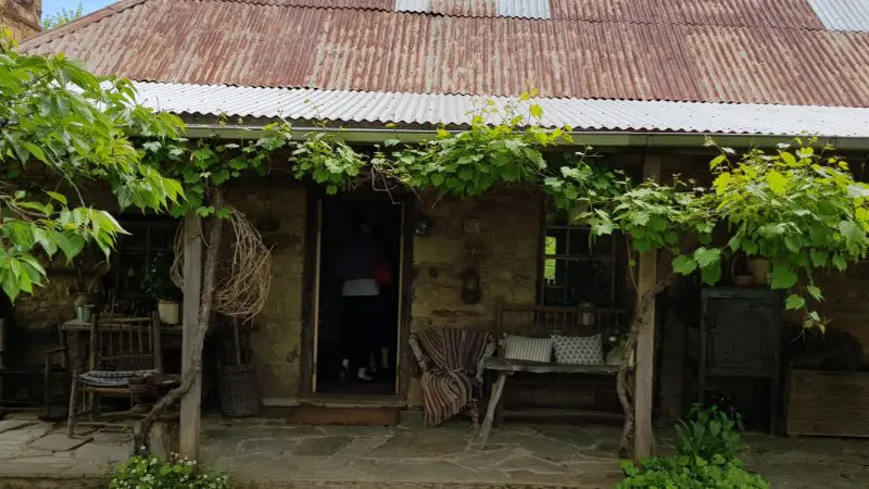 Quaint stone cottage with weathered metal roof, lush green vines, and a person entering—authentic Mt Macedon Ranges architectural charm.