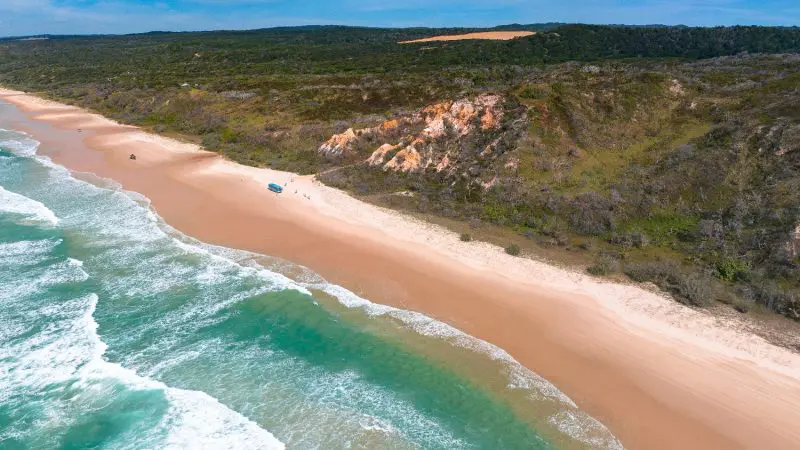 Stunning aerial view of Kgari 3 Day Getaway: pristine sandy beach, crystal-clear turquoise waters, lush green cliffs, and clear blue sky.
