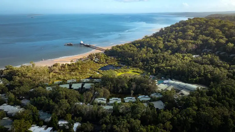Stunning aerial view of Kgari coastal resort by a lush forested hill, featuring a scenic pier stretching into vivid blue ocean waters.