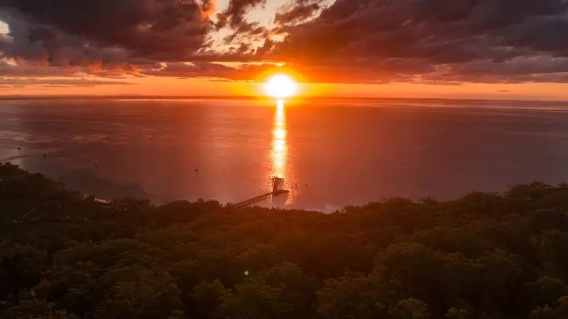 Breathtaking orange sunset over the ocean on a Kgari 3 Day Getaway, with a scenic pier and lush trees enhancing the peaceful view.