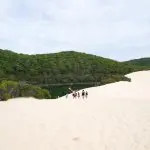 Tour group explores pristine white sand dunes with Island Expeditions K'gari Tours, approaching lush green lake under dramatic clouds.