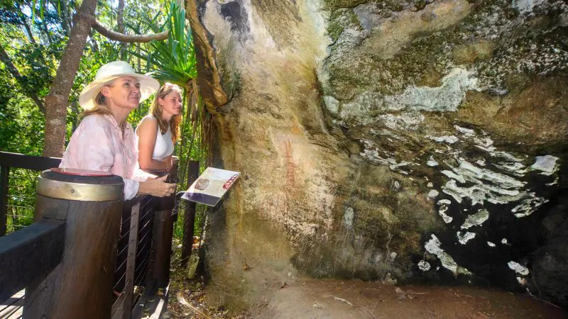 Two women viewing ancient rock art on a massive boulder during a Lady Enid Sailing tour in a vibrant forest, exploring local culture.